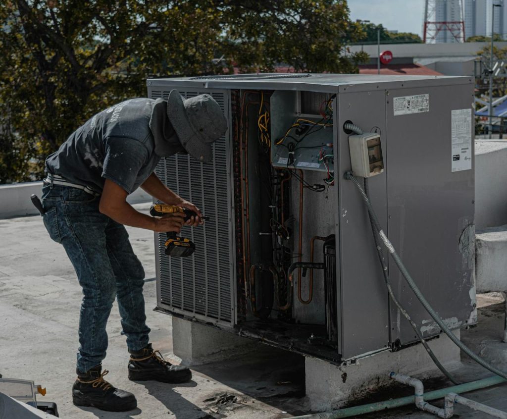 pexels-photo-5463587 A worker in a bucket hat repairs an outdoor air conditioning unit on a rooftop.